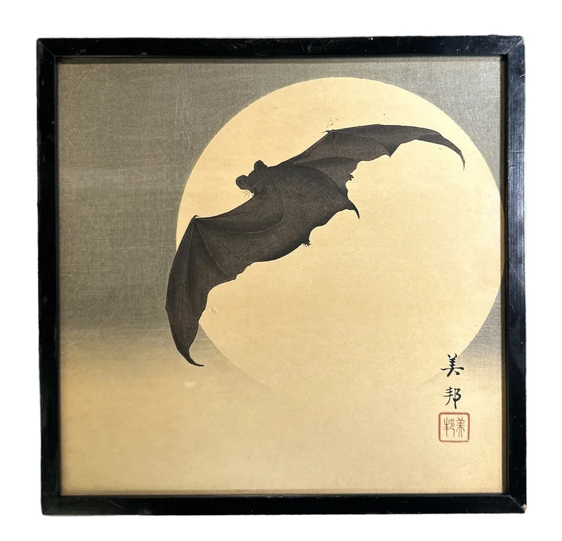A bat with spread wings crosses a large pale moon on a gold-toned ground, in a plain black frame, with the artist's signature and seal at the lower right.